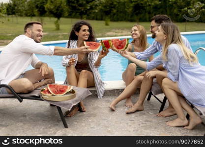 Group of young people sitting by the swimming pool and eating watermelon in the house backyard