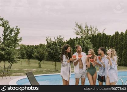 Group of young people standing by the swimming pool and eating watermellon in the house backyard