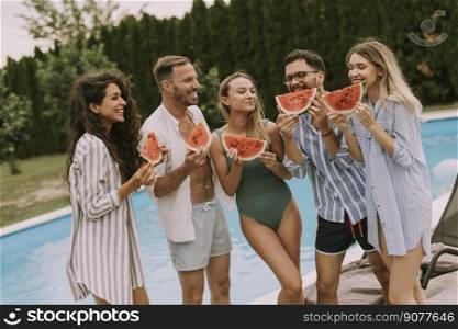 Group of young people standing by the swimming pool and eating watermelon in the house backyard