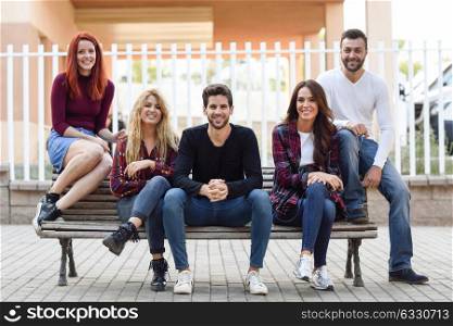 Group of young people together outdoors in urban background. Women and men sitting on a bench in the street wearing casual clothes.