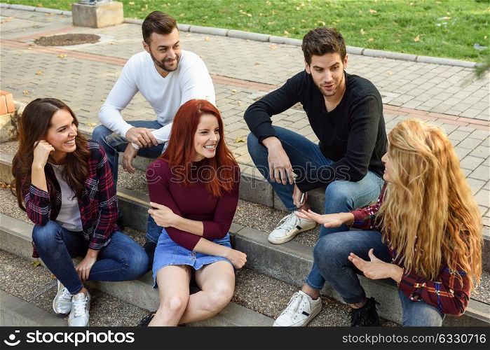 Group of young people together outdoors in urban background. Women and men sitting on stairs in the street wearing casual clothes.
