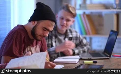 Group of young students studying together and preparing for exams at the table at home. Young hipster with beard writing notes in notebook concentratedly, his handsome blond friend working on laptop and making notes with pen in textbook. Side view.
