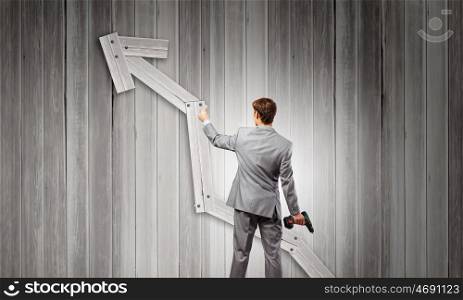 Growth concept. Rear view of businessman fixing wooden graph arrow with drill