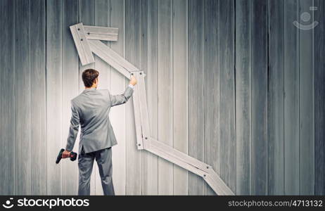 Growth concept. Rear view of businessman fixing wooden graph arrow with drill