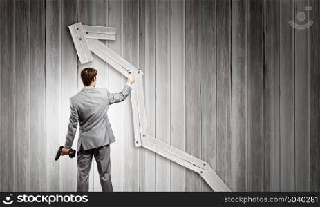 Growth concept. Rear view of businessman fixing wooden graph arrow with drill