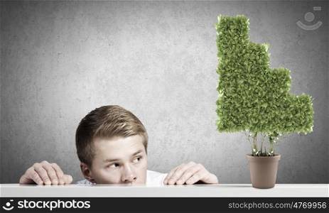 Growth concept. Young man looking out on tree in pot from under the table