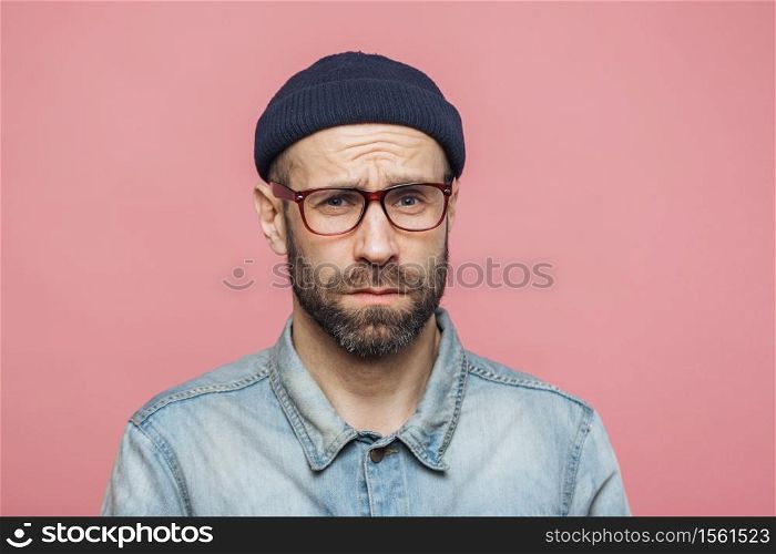 Grumpy bearded man with offended expression, being dissatisfied with something, frowns face, wears spectacles, hat and shirt, isolated over pink background. Displeasure and negativity concept