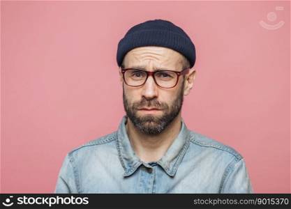 Grumpy bearded man with offended expression, being dissatisfied with something, frowns face, wears spectacles, hat and shirt, isolated over pink background. Displeasure and negativity concept