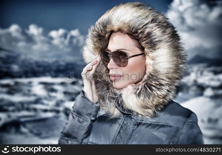 Grunge style photo of luxury woman portrait in wintertime outdoors, wearing stylish sunglasses and warm coat with furry hood and looking away, fashionable winter style concept