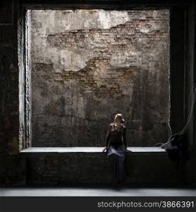 Grungy photo of lonely woman sitting at big window in old abandoned building