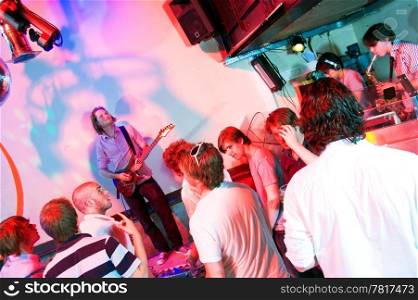 Guitarist on stage in a nightclub, with party crowd, saxophonist and DJ