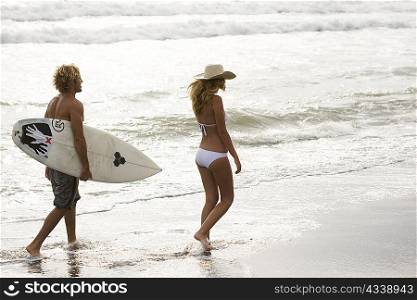 Guy and girl walking on beach in sunset, him with surf board
