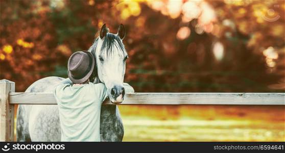 Guy bumped his head in neck of horse at fence in stable on background of autumn foliage