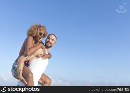 Guy carries his girlfriend piggy back style on the beach in the summer. They are laughing and smiling.