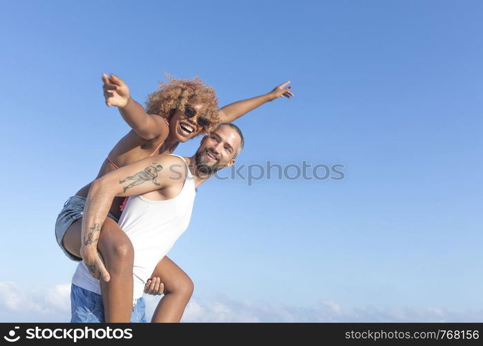 Guy carries his girlfriend piggy back style on the beach in the summer. They are laughing and smiling.