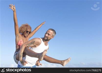 Guy carries his girlfriend piggy back style on the beach in the summer. They are laughing and smiling.