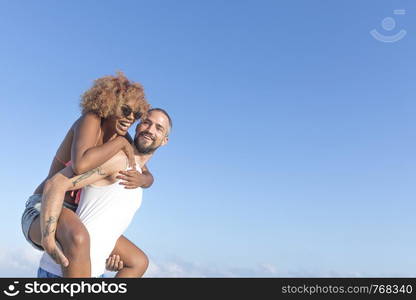 Guy carries his girlfriend piggy back style on the beach in the summer. They are laughing and smiling.