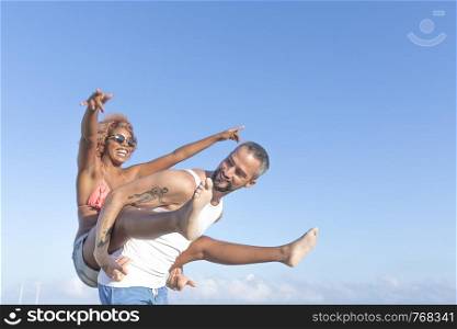 Guy carries his girlfriend piggy back style on the beach in the summer. They are laughing and smiling.