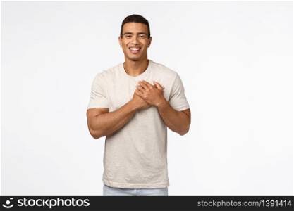 Guy feeling touched and flattered receiving praises. Charming hispanic young man in t-shirt, press hands to heart, smiling and laughing carefree, express gratitude, white background.. Guy feeling touched and flattered receiving praises. Charming hispanic young man in t-shirt, press hands to heart, smiling and laughing carefree, express gratitude, white background