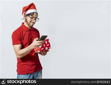 Guy in christmas hat holding gift box and smiling at smartphone. Christmas man holding gift box and telephone isolated. Handsome man in christmas hat holding gift and looking at phone