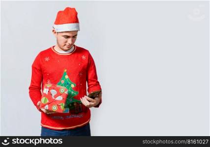 Guy in christmas hat holding gift box and smiling at smartphone. Christmas man holding gift box and telephone isolated. Handsome man in christmas hat holding gift and looking phone