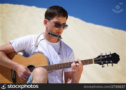 guy in sunglasses plays guitar and lip accordion sitting on sand
