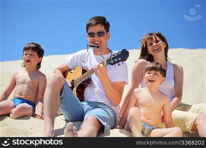 guy in sunglasses plays guitar and lip accordion with children and girl singing song sitting on sand