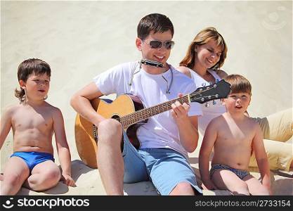 guy in sunglasses plays guitar and lip accordion with children and girl sitting on sand