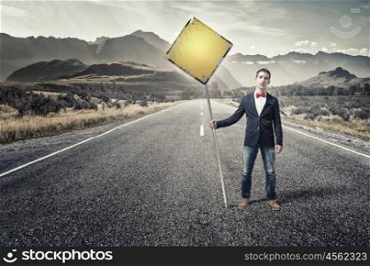 Guy showing roadsign. Young man in jacket and bowtie on road holding sign