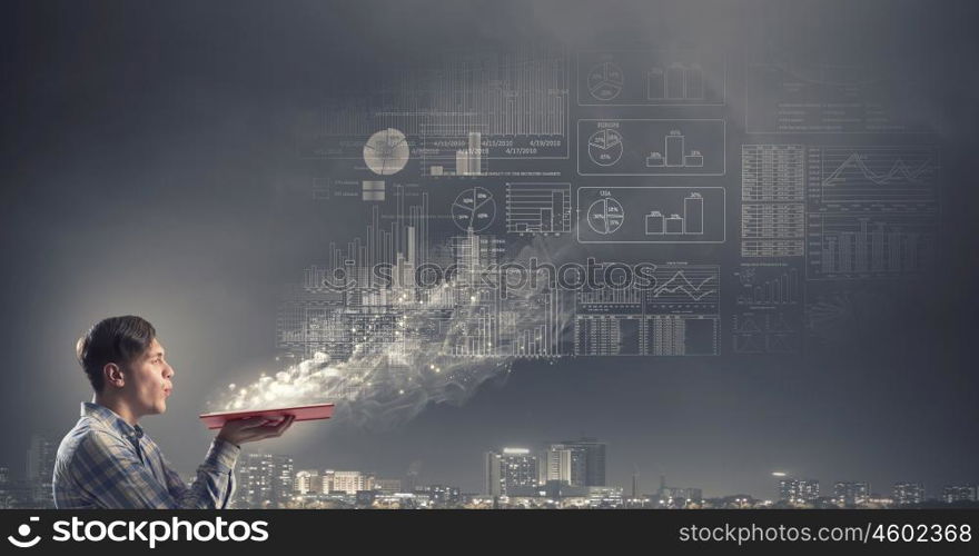 Guy studying finances. Young man with opened book in hands and infographs on wall blowing on pages