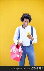 Guy with curly hair wearing shirt and suspenders. Black man with afro hairstyle carrying a sports bag and smartphone against a yellow urban background.. Black man with afro hairstyle carrying a sports bag and smartphone in yellow background.