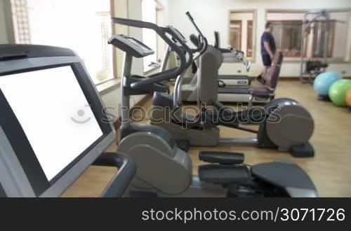 Gym with modern sport equipment. Blank screen of exercise machine in foreground, two men training in background