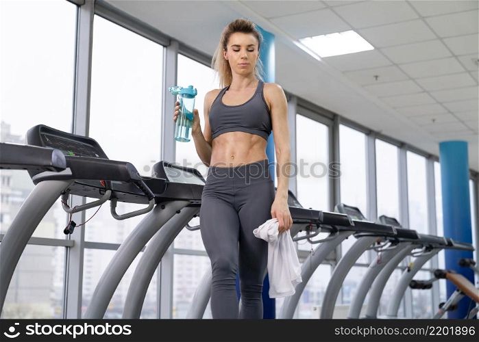 Gym woman working out drinking water standing by fitness machines inside in fitness center. Gym woman working out drinking water standing by fitness machines