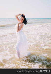 Gypsy young brunette girl in white maxi long dress standing on the waves in sea or ocean. Windy beach. Bohemian lifestyle. Portrait of happy sexy woman.