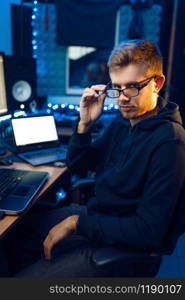 Hacker in hood at his workplace with laptop and desktop PC, website or corporate hacking, darknet user. Internet spy, crime lifestyle, risk job, network criminal. Hacker in hood at his workplace, corporate hacking