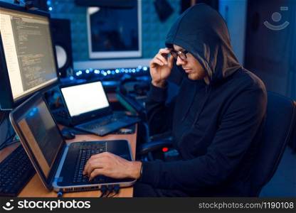 Hacker in hood at his workplace with laptop and desktop PC, website or corporate hacking, darknet user. Internet spy, crime lifestyle, risk job, network criminal. Hacker in hood at his workplace, corporate hacking
