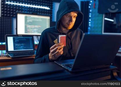 Hacker in hood shows bank credit card at his workplace with laptop and desktop PC, password or finance hacking, darknet user. Internet spy, crime lifestyle, risk job, network criminal. Hacker shows bank credit card, finance hacking