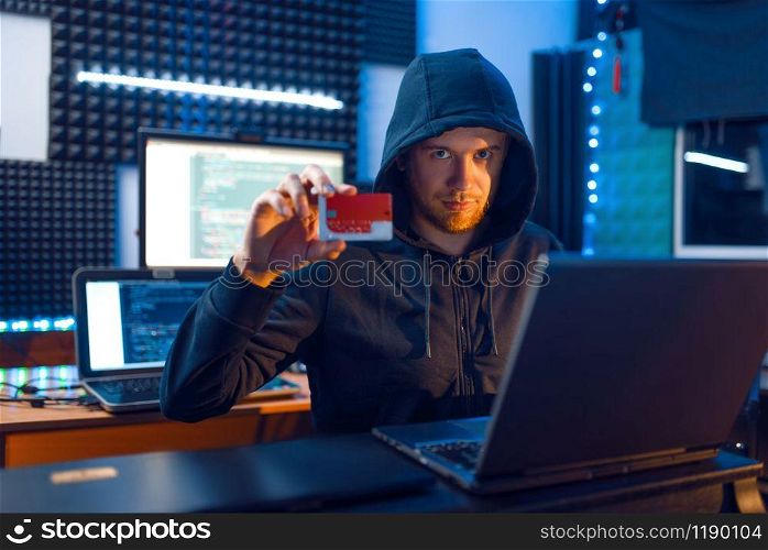 Hacker in hood shows bank credit card at his workplace with laptop and desktop PC, password or finance hacking, darknet user. Internet spy, crime lifestyle, risk job, network criminal