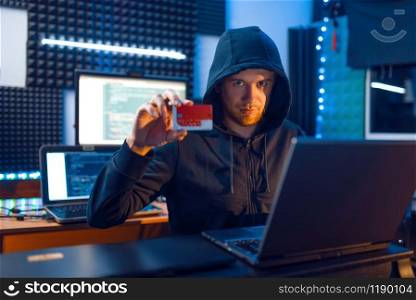 Hacker in hood shows bank credit card at his workplace with laptop and desktop PC, password or finance hacking, darknet user. Internet spy, crime lifestyle, risk job, network criminal
