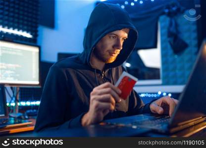 Hacker in hood shows bank credit card at his workplace with laptop and desktop PC, password or finance hacking, darknet user. Internet spy, crime lifestyle, risk job, network criminal. Hacker shows bank credit card, finance hacking