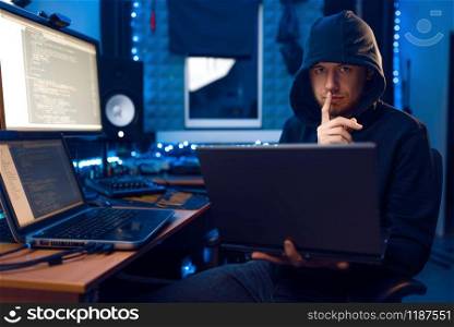 Hacker in hood shows thumbs up at his workplace with laptop and PC, password or account hacking. Internet spy, crime lifestyle. Hacker in hood shows thumbs up, network criminal