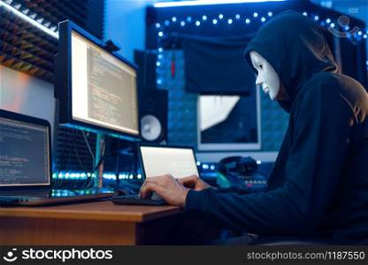 Hacker in mask and hood sitting at his workplace with laptop and PC, password or account hacking. Internet spy, illegal lifestyle, risk job, network criminal. Hacker in mask and hood, account hacking