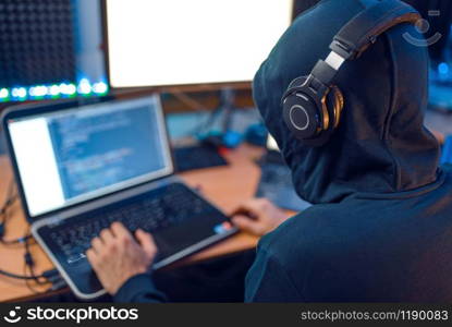 Hacker in the hood sitting at laptop, back view, information hacking. Internet spy, male programmer trying to hack an encrypted network. Hacker in the hood sitting at laptop, back view
