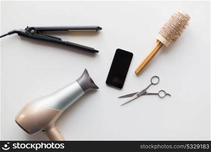 hair tools, beauty and hairdressing concept - smartphone, scissors, hairdryer with hot styling iron and curling brush on white background. smartphone, scissors, hairdryer, iron and brush