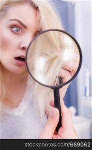 Haircare, bad effects of bleaching concept. Unhappy woman looking at ends of her blonde hair through magnifying glass. Woman looking at hair ends through magnifying glass