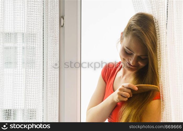 Haircare, morning routine concept. Woman sitting on windowsill brushing her long dark hair with comb.. Woman sitting on windowsill brushing her hair