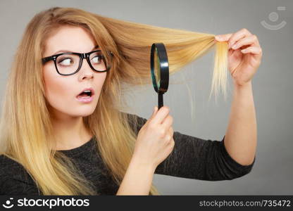 Haircare problems concept. Shocked blonde woman holding magnifying glass investigating her hair.. Woman holding magnifying glass looking at hair