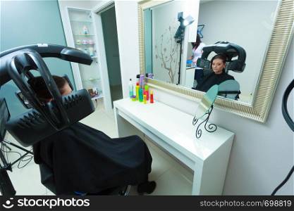 Haircare, relaxation and hairstyling concept. Woman sitting in black cape getting her hair dried under machine. Woman in hairdresser, drying hair under machine
