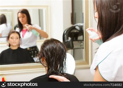 Haircare, relaxation and hairstyling concept. Woman sitting in black cape getting her hair sprayed with conditioner by lady hairdresser in beauty salon. Hairdresser spraying conditioner on woman hair