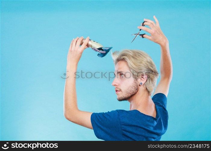 Hairdo changing hairstyle concept. Cool male hairstylist making self trendy haircut, fashion blonde metrosexual model going to shave his long hair, using shaver and scissors. Man going to shave his long hair